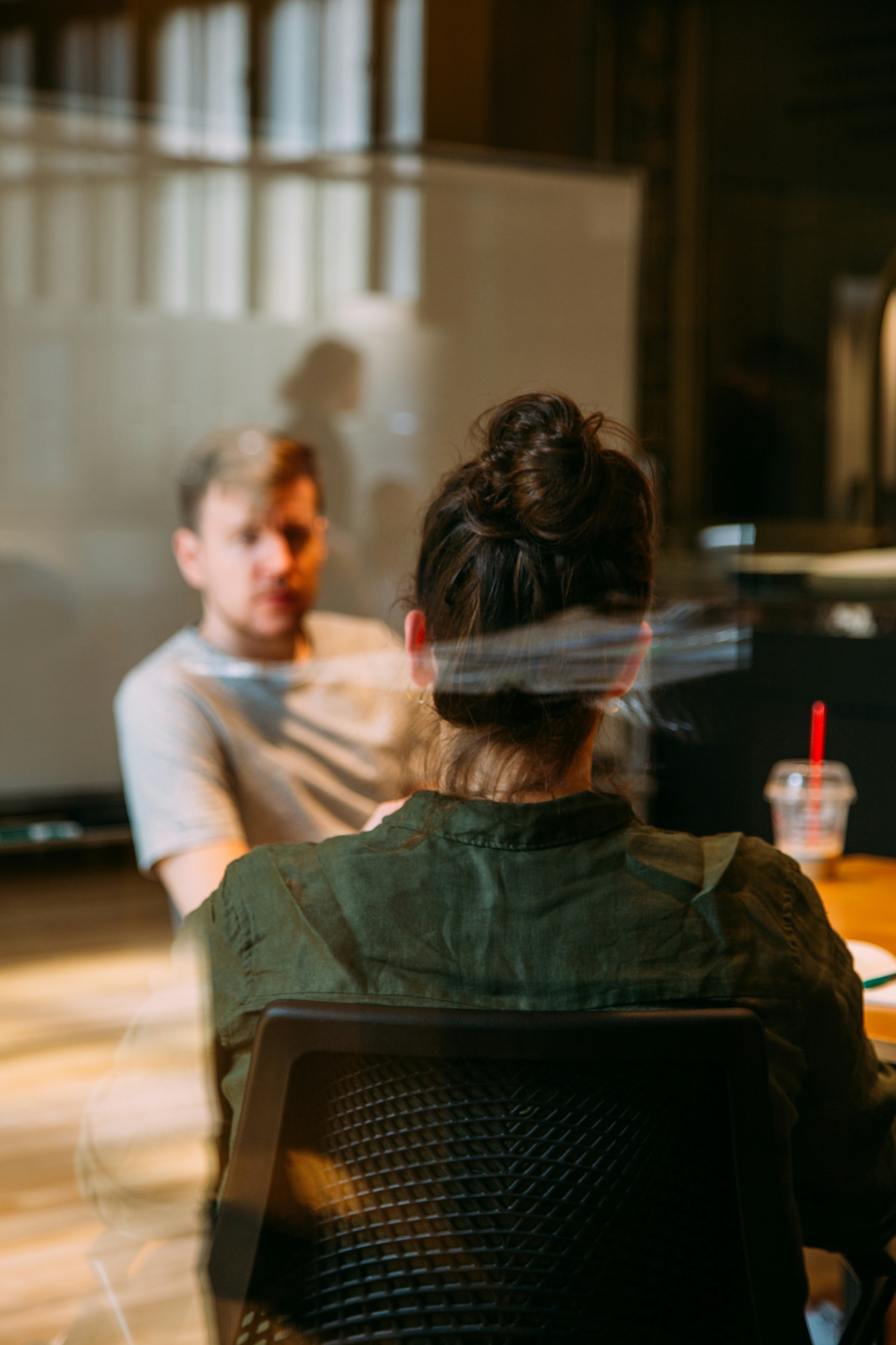 Two people having a conversation indoors, seen through a glass panel with reflections.