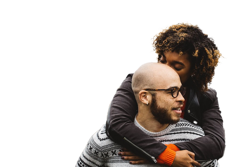 Woman with curly hair hugging and kissing the head of a smiling man wearing glasses and a patterned sweater outdoors.